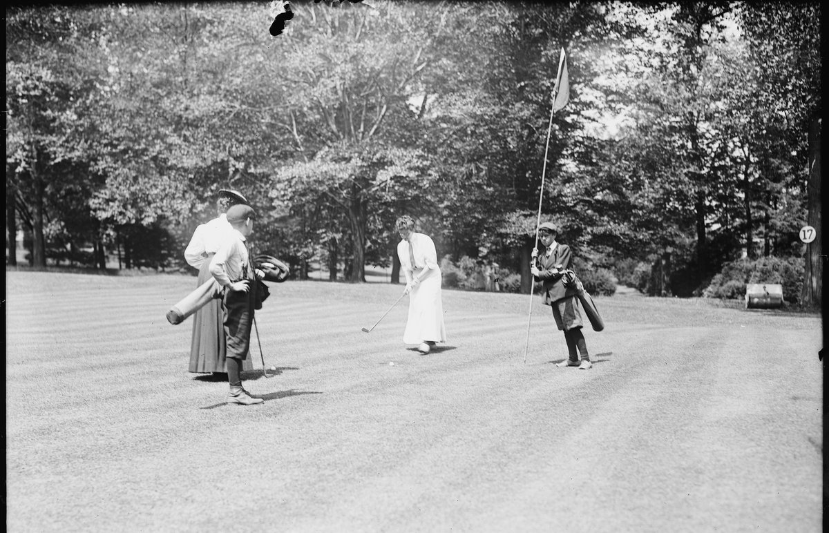 Photographie ancienne de deux joueuses et d'un caddie sur un green, drapeau au fond